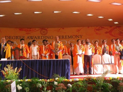 A row of academics in orange and saffron gowns stands behind a blue-draped table on a stage, beneath a banner reading Degree Awarding Ceremony.