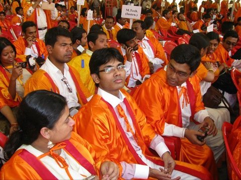 Rows of graduates in orange gowns sit on red plastic chairs under a pale tent, several looking toward the camera; a sign reads M.Tech.