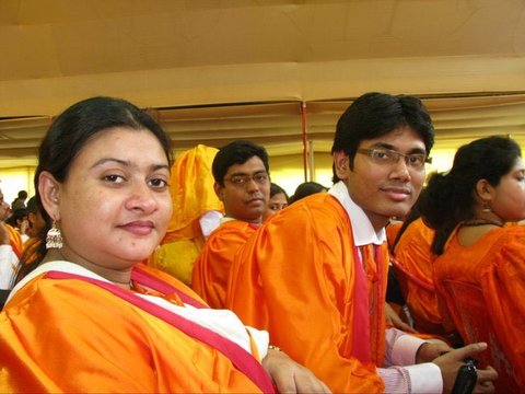 Two graduates in orange gowns turn to face the camera from their seats beneath a tent, others in gowns around them.