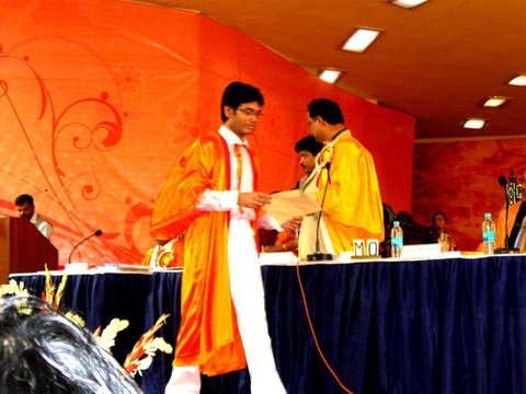 A graduate in an orange gown reaches across a stage table to receive a document from a man in a yellow and orange gown, against a patterned orange backdrop.