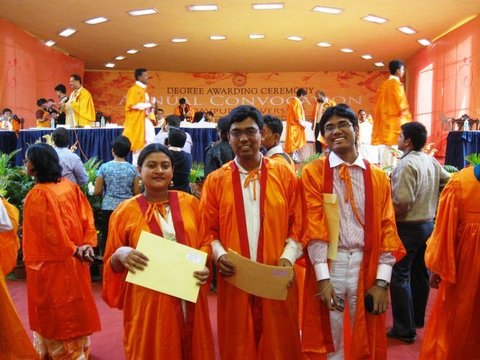 Three graduates in orange gowns hold yellow envelopes in a crowded hall, the Annual Convocation banner of Jadavpur University behind them.