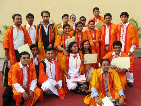About twenty graduates in orange gowns pose in three rows on a red carpet before a cream wall, several holding yellow envelopes.
