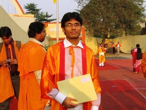 A graduate in an orange gown holds a yellow envelope on a red carpet, a tall yellow flag and a bare tree against a pale sky behind him.