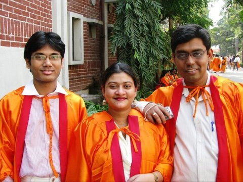 Three graduates in orange gowns with red sashes smile beside a red-brick wall, green foliage and more gowned figures behind them.
