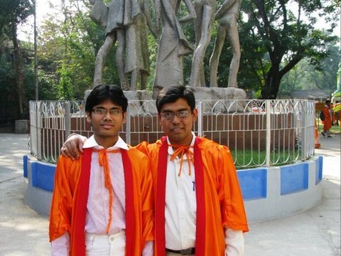 Two graduates in orange gowns stand arm in arm in front of a grey stone sculpture of striding figures ringed by a white railing.