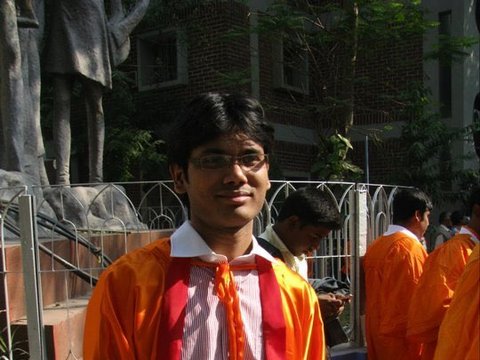 A graduate in an orange gown over a striped shirt and white trousers stands in bright sun, a stone statue and railing behind him.