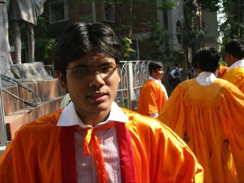 Close view of a graduate in an orange gown and glasses, sunlit, with a stone statue, railing and other gowned figures behind.