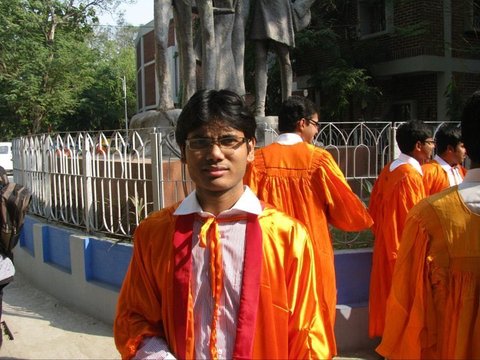 A graduate in an orange gown faces the camera while others in gowns stand with their backs turned at a railed statue plinth under trees.