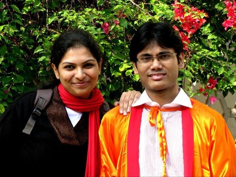 A woman in a black top and red scarf and a graduate in an orange gown smile in front of a hedge of magenta bougainvillea.
