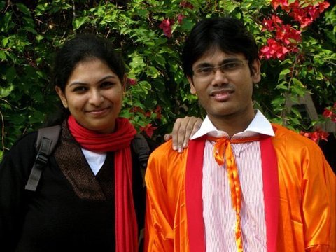 A woman with a red scarf and a graduate in an orange gown and white trousers stand before green leaves and magenta bougainvillea.