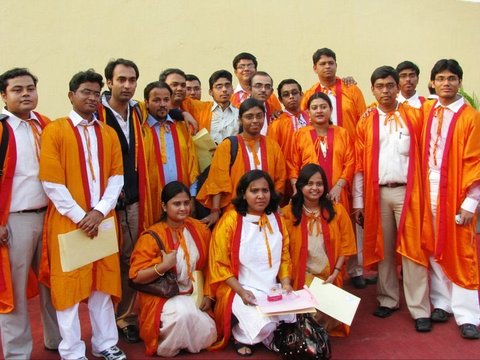 A large group of graduates in orange gowns with red and white sashes, some holding yellow folders, gathered on a red carpet before a pale wall.