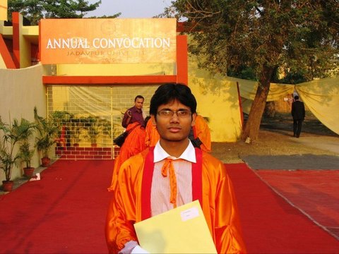 A graduate in an orange gown holding a yellow folder on a red carpet, an Annual Convocation banner and a gate behind him.