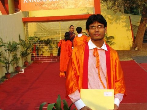A graduate in an orange gown stands on a red carpet holding a yellow folder, with the Annual Convocation banner of Jadavpur University above the gate behind.