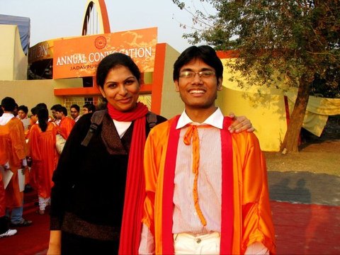 A smiling graduate in an orange gown beside a woman in a red scarf and black jacket, graduates and a convocation banner in the background.