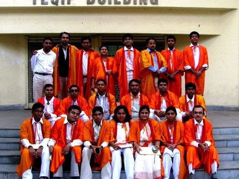 Three rows of graduates in orange gowns posed on the steps of a building whose facade reads TEQIP BUILDING.