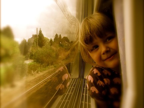 A young child in an elephant-print top leans against a train window, smiling as trees and houses slide past in warm, hazy light.