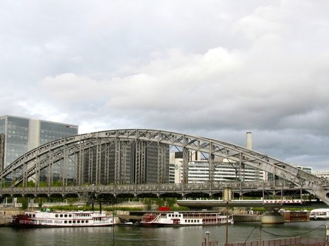 A great riveted steel arch carries the metro viaduct over the Seine, with excursion boats moored below and office blocks behind.