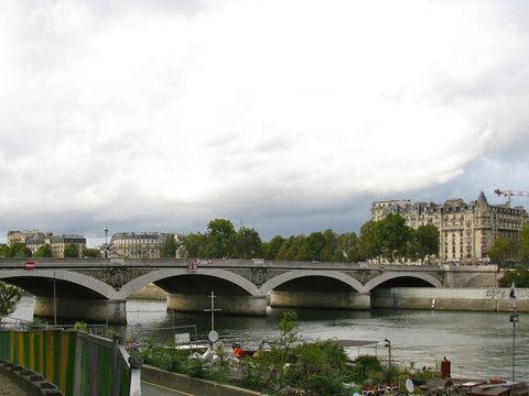 A five-arched stone bridge spans a wide grey river beneath an overcast sky, with Haussmann-style apartment blocks and a crane on the far bank.