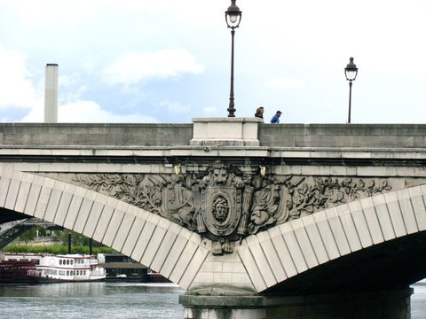 A carved stone cartouche with a lion's mask and a wreathed face sits at the crown of a bridge arch, two people passing beneath its lamps.