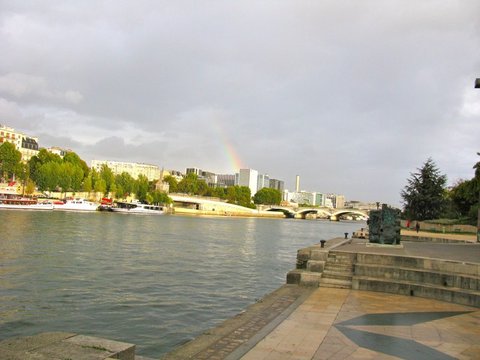 A faint rainbow drops into a cityscape of mid-rise buildings across a wide river, moored boats along the far bank and wet paving in the foreground.