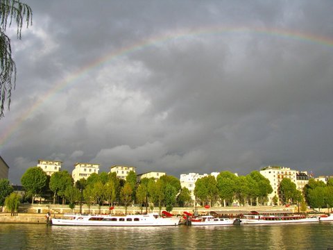 A broad rainbow spans a grey sky over sunlit cream-coloured apartment blocks, plane trees and white river boats, a willow branch framing the left edge.
