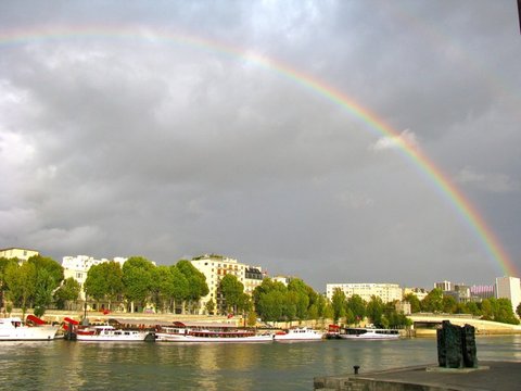 A rainbow curves from the top left down toward tower blocks on the far bank, with a line of moored cruise boats and bronze sculptures on the quay.