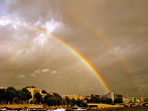 Two rainbows, one bright and one faint, arc across a brooding sky above a riverside skyline, a stone bridge and a row of moored boats.