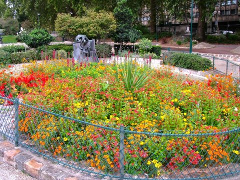 A bed of orange, red and yellow bedding plants around a spiky yucca, edged by a low green wire fence, with a bronze sculpture and trees behind.