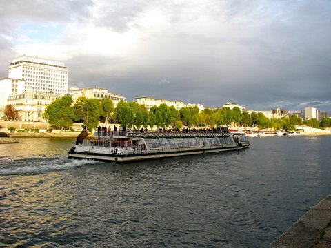 A long glass-roofed excursion boat crowded with passengers churns along a wide river in low golden light, tall buildings on the far bank.