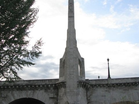 A tall stone pylon topped by a veiled figure rises from the end of a bridge beside the Seine, walkers passing under its arch beneath a cloudy sky.