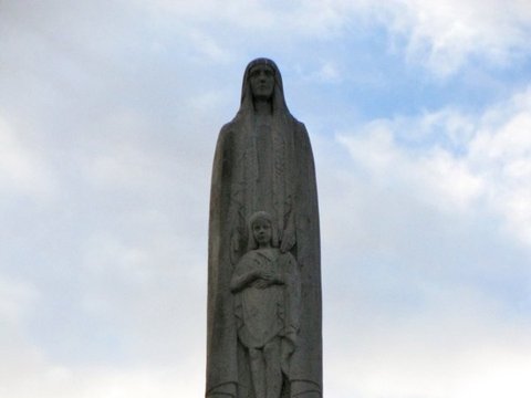 Close view of a stone statue of a veiled woman sheltering a child, set on a battlemented pylon against a bright, broken sky.