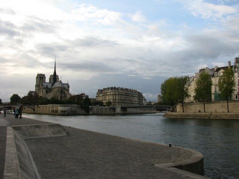 Wide view down the Seine from a cobbled quay, the spire and flying buttresses of Notre-Dame rising above the trees of the Île de la Cité.