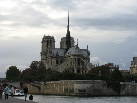 The towers, spire and flying buttresses of Notre-Dame seen from across the Seine, a few people on the quay below a grey sky.