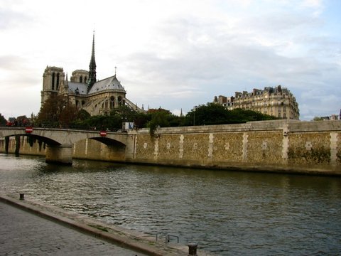 A stone bridge and long quay wall along the Seine, with the spire and towers of Notre-Dame above the trees on the left.
