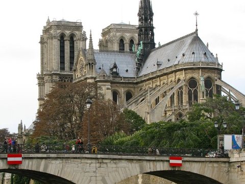 The spire, towers and buttressed apse of Notre-Dame rise above a two-arched stone bridge with red-and-white markers over the Seine.