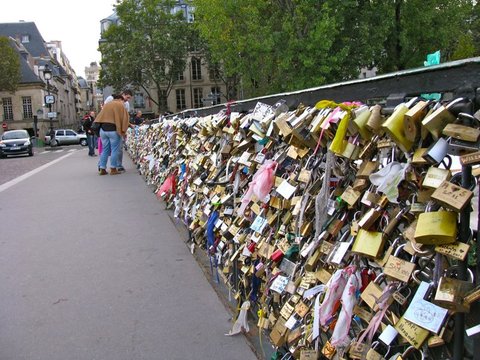 A bridge railing densely covered in brass padlocks and ribbons stretches toward tall stone buildings, two people pausing to look.