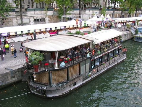 Seen from above, diners sit beneath the canopy of a moored restaurant barge while market stalls and white tents line the quay behind.