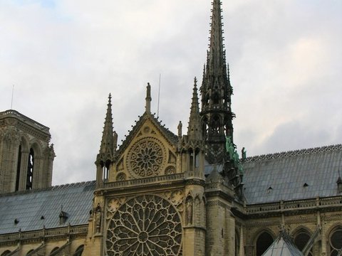 Close view of the south rose window and gabled transept of Notre-Dame, the dark lead spire climbing into a pale cloudy sky.