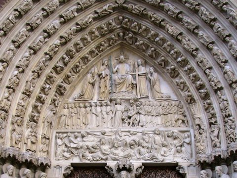 A pointed stone portal crowded with carved figures in its arches and tympanum, dark ironwork doors below and four visitors at the foot.