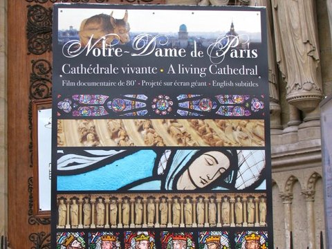 A black poster for a film about Notre-Dame de Paris, printed with stained-glass and sculpture details, stands before a carved doorway.