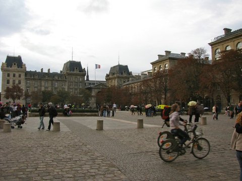 Cyclists ride across a wide cobbled square toward mansard-roofed buildings and a tricolour flag, visitors scattered under an overcast sky.