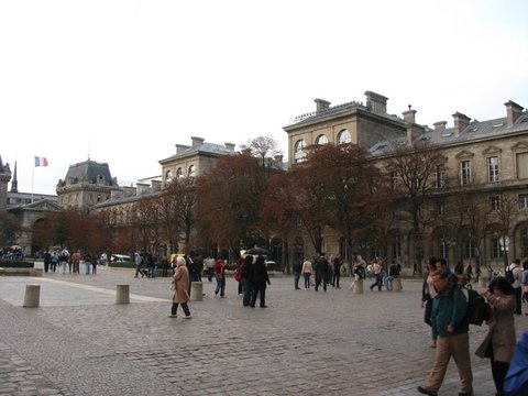 Visitors cross a wide cobbled square under a grey sky, with autumn trees and a long mansard-roofed stone facade flying a French flag behind them.