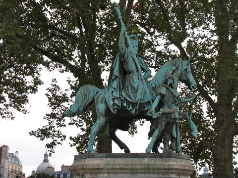 Green bronze of a crowned rider raising a sceptre on horseback, flanked by two armoured attendants, framed by plane-tree branches against a white sky.