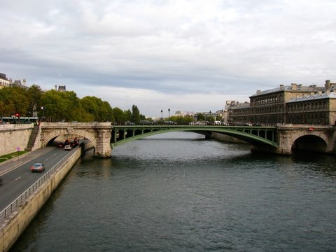 A single green iron arch spans the grey Seine between stone abutments, traffic passing under the quay arches beneath a cloudy sky.