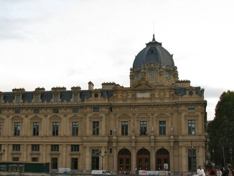 Ornate stone courthouse with a slate dome and rows of tall windows under a flat white sky, a green hoarding and parked vans along its base.