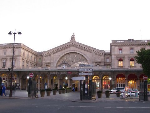 The arched glass facade of Gare de l'Est with its rooftop statue and clock, lamps and awnings lit as the sky fades to pale blue.