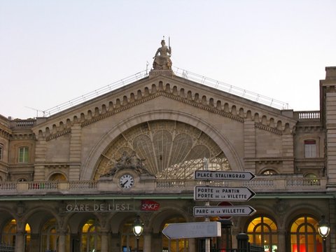 Fan-shaped iron tracery and a clock beneath the sculpted pediment of Gare de l'Est, with street signs pointing to Place Stalingrad and Porte de Pantin.