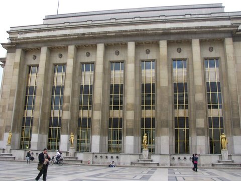Fluted stone pillars and tall gold-framed windows of the Palais de Chaillot, gilded figures standing between them above a patterned pavement.