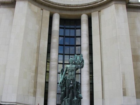 Curved limestone wall of the Palais de Chaillot carrying gold lettering above a bronze figure holding a lyre, with two people and a pram at its foot.