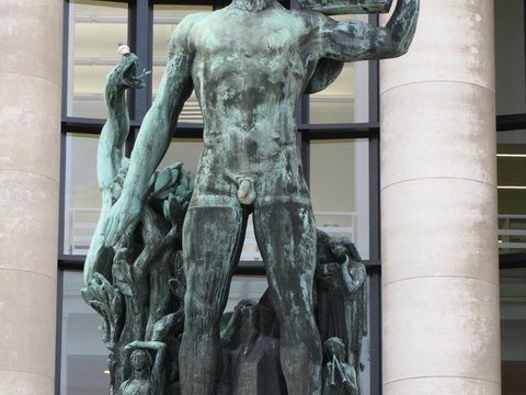 Green-patinated bronze of a standing figure holding a lyre aloft, set between pale stone columns in front of tall gridded windows.